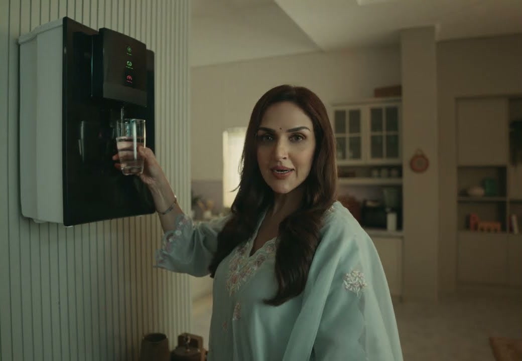 A woman in a pale blue kurta fills a glass at a wall-mounted water dispenser