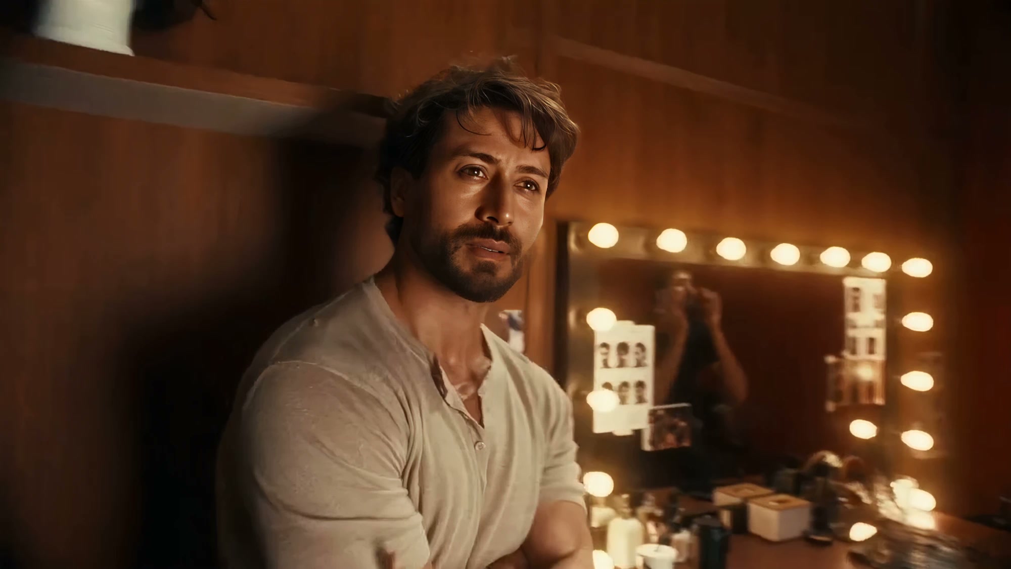 A man leans against a wall in a warmly lit backstage dressing room, mirror lights behind him